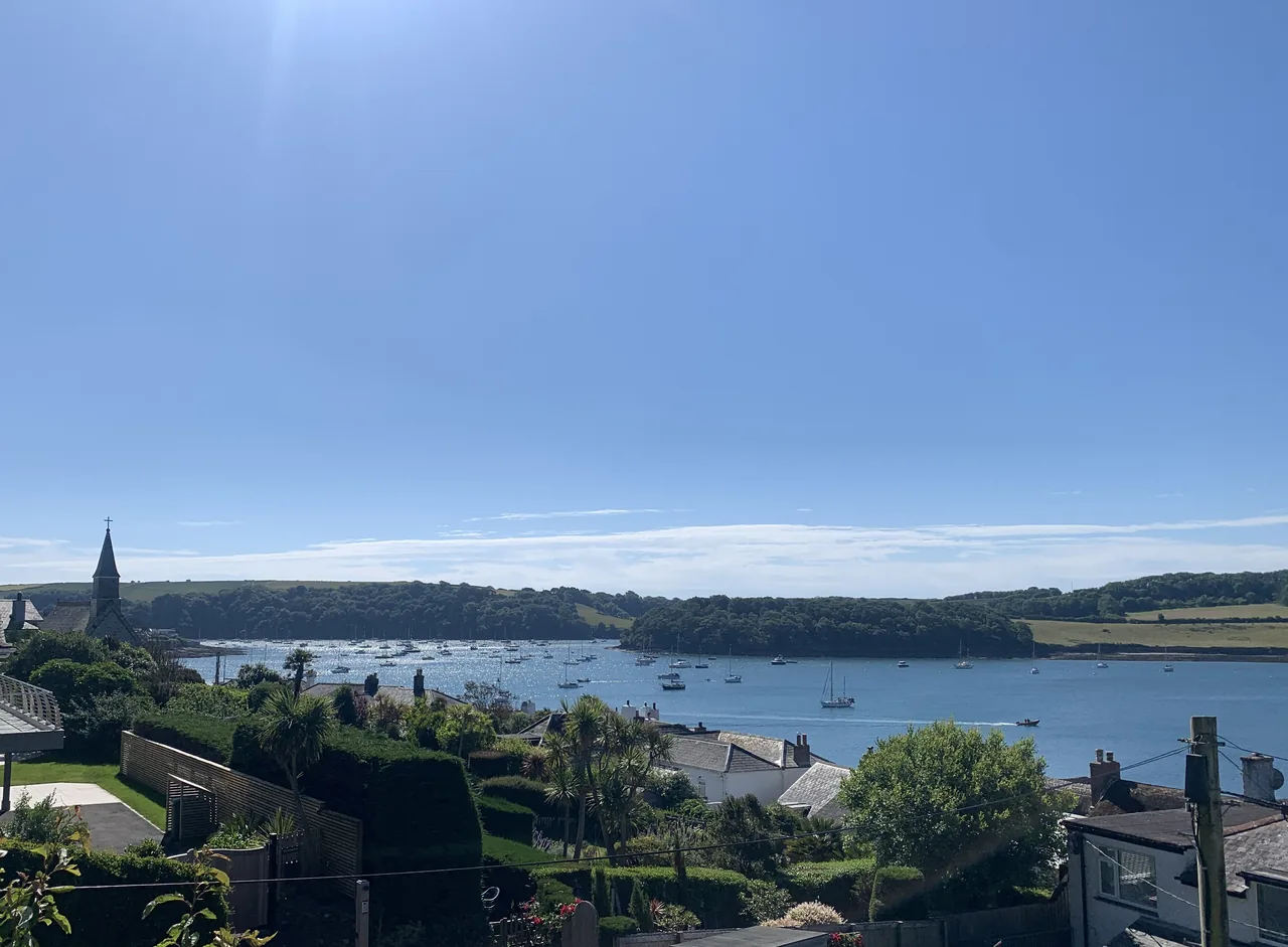 Rooftops and a church spire in the foreground, with moored boats and a wooded shoreline across the water.